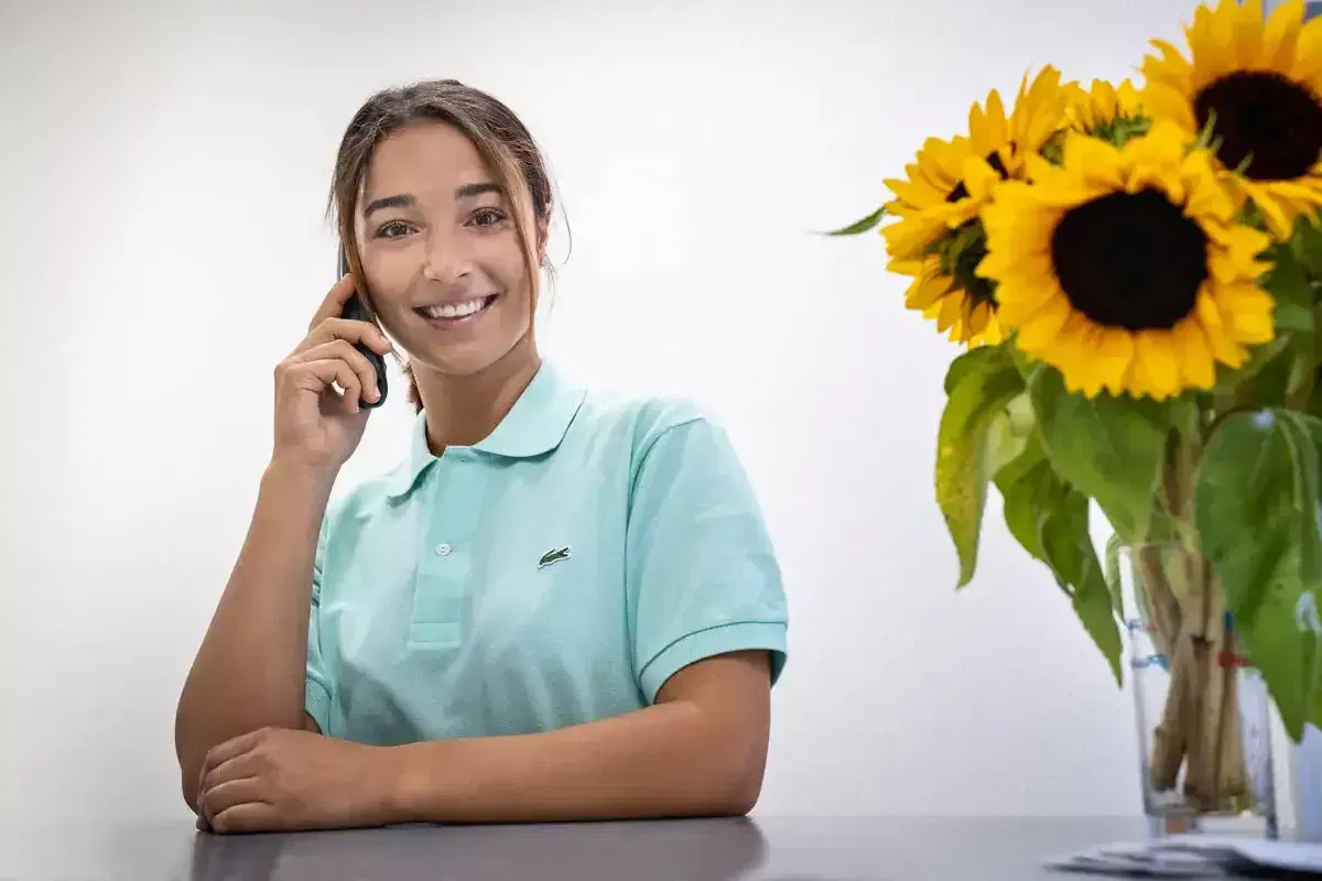 Frau in mintgrünem Polohemd telefoniert lächelnd, neben einem Strauß Sonnenblumen auf einem Tisch.